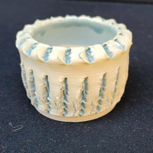 May include: A small white ceramic planter with a blue glaze. The planter has a decorative design of vertical lines and a scalloped rim.