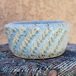 Może przedstawiać: Teksturowana, jasnoniebieska ceramiczna miska z szorstkim, geometrycznym wzorem. Miska ma szeroki, otwarty wierzch i lekko zaokrągloną podstawę. Zewnętrzna strona ma postarzany wygląd, sugerujący jakość rzemieślniczą. Miska stoi na wzorzystej tkaninie.