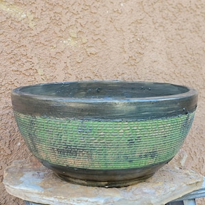 May include: A large, round ceramic bowl with a textured green and black woven pattern around the middle. The rim and base are dark gray. The bowl is sitting on a stone surface.