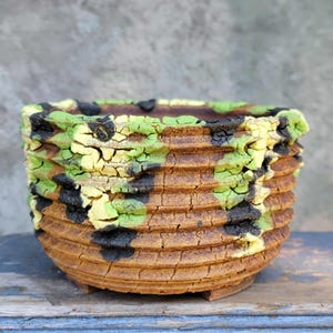 May include: A brown ceramic pot with a textured surface. The pot is decorated with black, green, and yellow paint in a camouflage pattern.