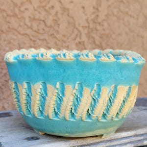 May include: A turquoise ceramic bowl with a textured, carved design. The rim and carved details are a light beige color. The bowl is sitting on a wooden surface.