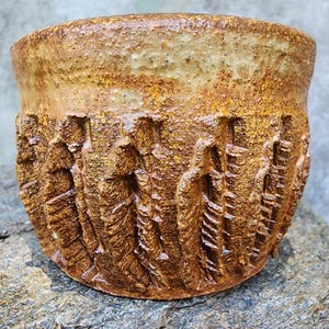 May include: A handcrafted ceramic bowl with a textured, earthy design. The bowl features a rough, carved pattern around the base and a speckled, light brown and yellow glaze. The bowl is sitting on a gray stone surface.