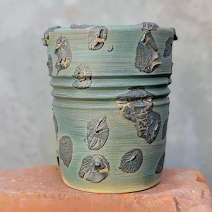May include: A handmade ceramic planter with a textured surface and a unique design. The planter is cylindrical, with a light teal base color and dark gray, textured, organic shapes. The planter has horizontal ridges and a yellow base.