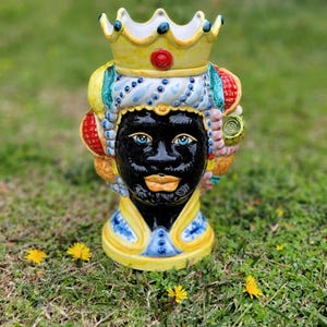 May include: A ceramic planter shaped like a black face with a yellow crown, blue and white turban, and colorful fruit and flower accents. The planter is sitting on a bed of green grass.