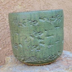 Puede incluir: Una maceta de cerámica verde hecha a mano con una superficie texturizada y rugosa. La maceta cilíndrica tiene una abertura ancha y una base ligeramente redondeada. El exterior presenta un esmalte verde terroso moteado con motas más oscuras, lo que le da un aspecto rústico. Ideal para uso en interiores o exteriores.