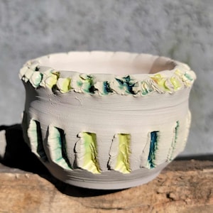 May include: A handmade ceramic bowl with a white exterior and a textured rim. The bowl features vertical cut-out designs with yellow and blue accents. The rim is decorated with small, colorful floral-like details. The bowl is set on a wooden surface.