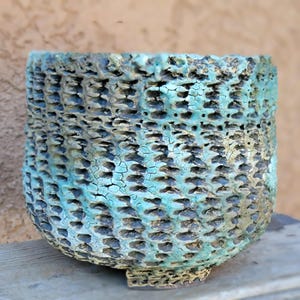 May include: A handcrafted ceramic pot with a textured, basket-weave design. The pot features a turquoise and black color scheme with a distressed, crackled finish. The unique vessel is suitable for plants or decorative use.
