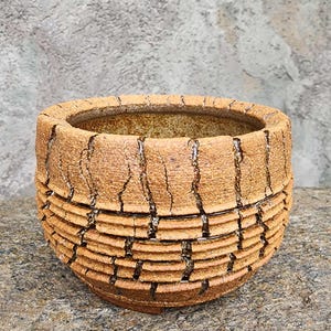 May include: A round ceramic planter with a textured, tan-colored exterior featuring deep, vertical fissures and horizontal ridges. The surface has a rough, earthy appearance with dark brown glaze accents within the cracks and a wide, open top.
