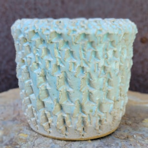 May include: A small, handmade ceramic pot with a textured surface. The pot is light blue with a pattern of raised, star-shaped designs. The rim is slightly uneven, and the base is a light tan color. This decorative pot is suitable for small plants.