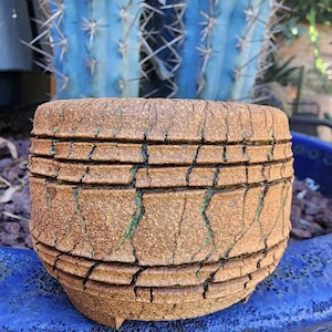 May include: A cylindrical ceramic planter with a textured, light brown, crackled surface finish. The exterior features horizontal ridges and deep vertical fissures that reveal a green hue within the cracks. The planter has a flat base with small feet.