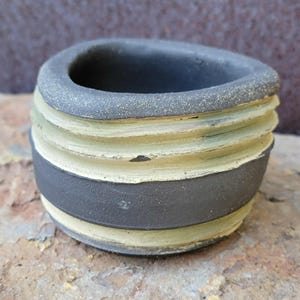 May include: A small, handcrafted ceramic pot with a dark gray rim and base, and alternating bands of light green and dark gray. The pot has a textured, ridged design and sits on a rough, brown surface.