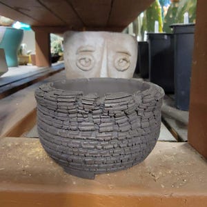 May include: A dark gray, textured ceramic pot with a rough, layered design. The pot has a wide, open top and a slightly rounded shape. The pot is sitting on a wooden shelf, with other pots and plants visible in the background. A decorative face pot is in the background.