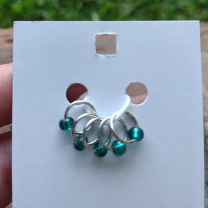 May include: Set of six teal colored stitch markers with silver rings. The markers are displayed on a white card.