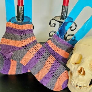 May include: A pair of hand-knitted socks with a striped pattern in orange, purple, and gray. The socks have a lace-like design on the upper part and are displayed on a black surface.