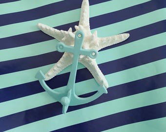 Può includere: Una stella marina bianca e un'ancora azzurra sono disposte su uno sfondo a righe blu e azzurre. L'ancora è posizionata sopra la stella marina, creando un tema nautico.
