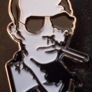 May include: A black and white enamel pin of a man wearing sunglasses and smoking a cigarette. The man has a serious expression on his face.