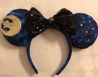Puede incluir: Un par de orejas de ratón azules y doradas con un lazo negro de lentejuelas. Las orejas tienen un estampado de galaxia con estrellas y un símbolo dorado de la Alianza Rebelde.