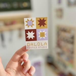 May include: A rectangular sticker featuring a patchwork design with star-shaped and square patterns in shades of purple, gold, and brown. The text "DALOLA TEXTILE STUDIO" is printed in a serif font below the design.