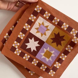 Op de afbeelding: Twee vierkante, roestkleurige bandana's met een geometrisch sterrenpatroon. Elke bandana heeft een centraal ontwerp van vier vierkanten, elk met een ster in wit, bruin, goud en lavendel. De rand heeft een herhalend patroon van kleine vierkanten.