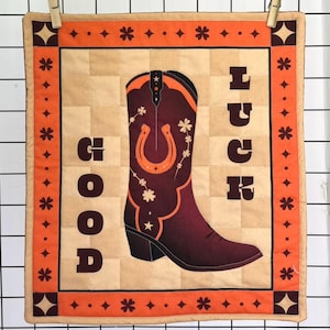 Peut inclure: Un carré matelassé avec un fond beige et une bordure orange. Le motif présente une botte de cowboy bordeaux avec un fer à cheval et des motifs floraux. Les mots "GOOD LUCK" sont imprimés verticalement. Le carré est maintenu par deux pinces à linge.