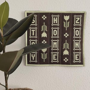Könnte beinhalten: Ein quadratisches, braunes Wandbehang mit dem Wort „HOME“ in weißen Blockbuchstaben. Die Buchstaben sind in vertikalen Spalten angeordnet, getrennt durch stilisierte Blumen- und geometrische Designs. Der Rand ist hellgrün.