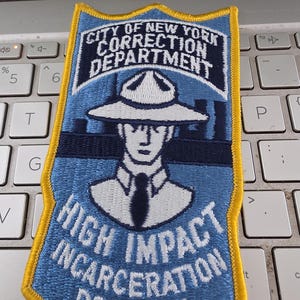 May include: A shield-shaped embroidered patch with a yellow border. The light blue background features a dark blue silhouette of a person wearing a wide-brimmed hat and tie. Text reads CITY OF NEW YORK CORRECTION DEPARTMENT and HIGH IMPACT INCARCERATION PROGRAM.