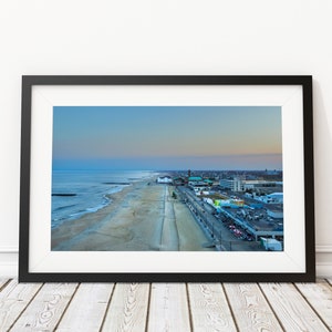 May include: An aerial view of a beach town at sunset. The beach is sandy and the sea is blue. There are buildings and a boardwalk in the distance.