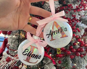 Può includere: Due palline di Natale trasparenti con glitter, con i nomi "Amelia" ed "Eliana" stampati sopra. Ogni ornamento ha un nastro e un fiocco rosa. Lo sfondo include un albero di Natale con bacche rosse.