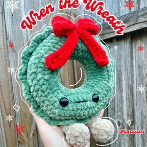 May include: A green crocheted wreath with a red bow and two small legs. The wreath has a cute cartoon face with black eyes. The text "Wren the Wreath" is written in a whimsical font above the wreath.