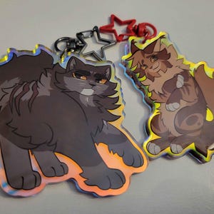 May include: Two iridescent keychains featuring cartoon cats. The first keychain features a black cat with a white chest and a white stripe on its back. The second keychain features a brown cat with a white chest and a white stripe on its back.