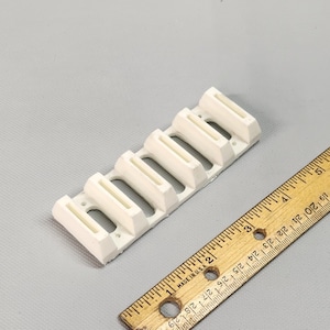 Könnte beinhalten: Weiße Wandhalterung aus Kunststoff mit sechs Schlitzen zur Organisation von Gegenständen. Die Halterung ist etwa 10 cm lang.
