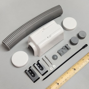 Puede incluir: Un surtido de componentes de plástico blanco y gris, que incluyen una manguera corrugada, una carcasa rectangular, dos tapas redondas y varias piezas pequeñas. También hay una regla de madera con marcas en pulgadas.