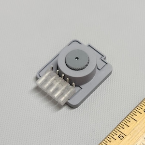 May include: A gray electronic component with a circular dial and a clear plastic connector. The component is rectangular with a raised circular section. A wooden ruler is in the lower right corner.