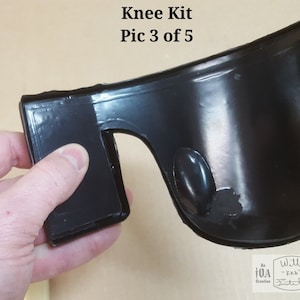 Pode incluir: Close-up de uma joelheira preta com uma superf&iacute;cie lisa e brilhante. A joelheira tem uma borda grossa e arredondada e um pequeno furo redondo no lado. A joelheira est&aacute; sendo segurada por uma m&atilde;o.