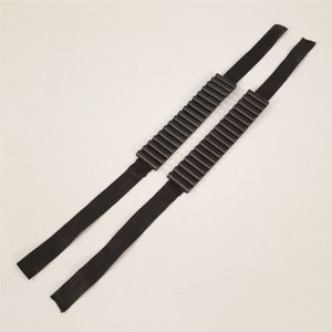 以下が含まれることがあります： 黒いショルダー ストラップ 2 本。等間隔に配置された、溝のある黒いプラスチック製の部品が並んでいます。 ストラップは織られた耐久性のある素材で作られています。 背景は無地のオフホワイトです。