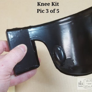 May include: Close-up of a black knee pad with a smooth, shiny surface. The knee pad has a thick, rounded edge and a small, round hole on the side. The knee pad is being held by a hand.