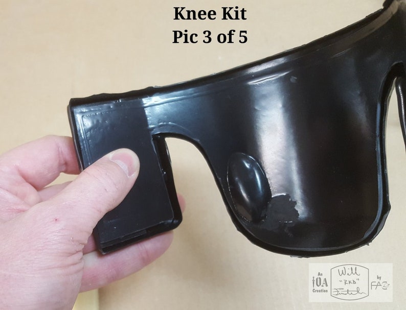 May include: Close-up of a black knee pad with a smooth, shiny surface. The knee pad has a thick, rounded edge and a small, round hole on the side. The knee pad is being held by a hand.