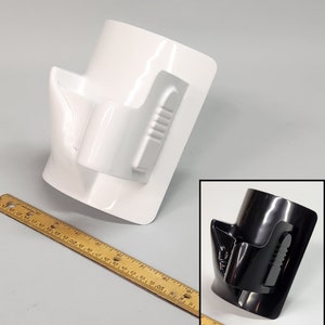 May include: A white plastic holder with a black plastic insert. The holder has a curved shape and a slot for a phone or other device. The insert has a textured surface and a raised lip.