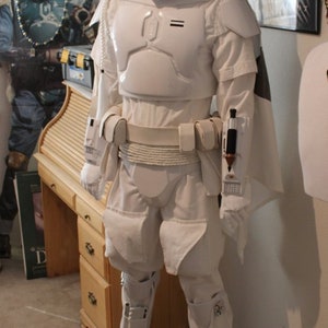 May include: A white Boba Fett costume with a white helmet and a white cape. The costume is standing on a brown carpet in front of a wooden desk. The costume is wearing a white belt with a rope detail. The costume has a white chest piece with a black visor. The costume has a white cape with a black trim. The costume has a white belt with a rope detail. The costume has a white chest piece with a black visor. The costume has a white cape with a black trim.