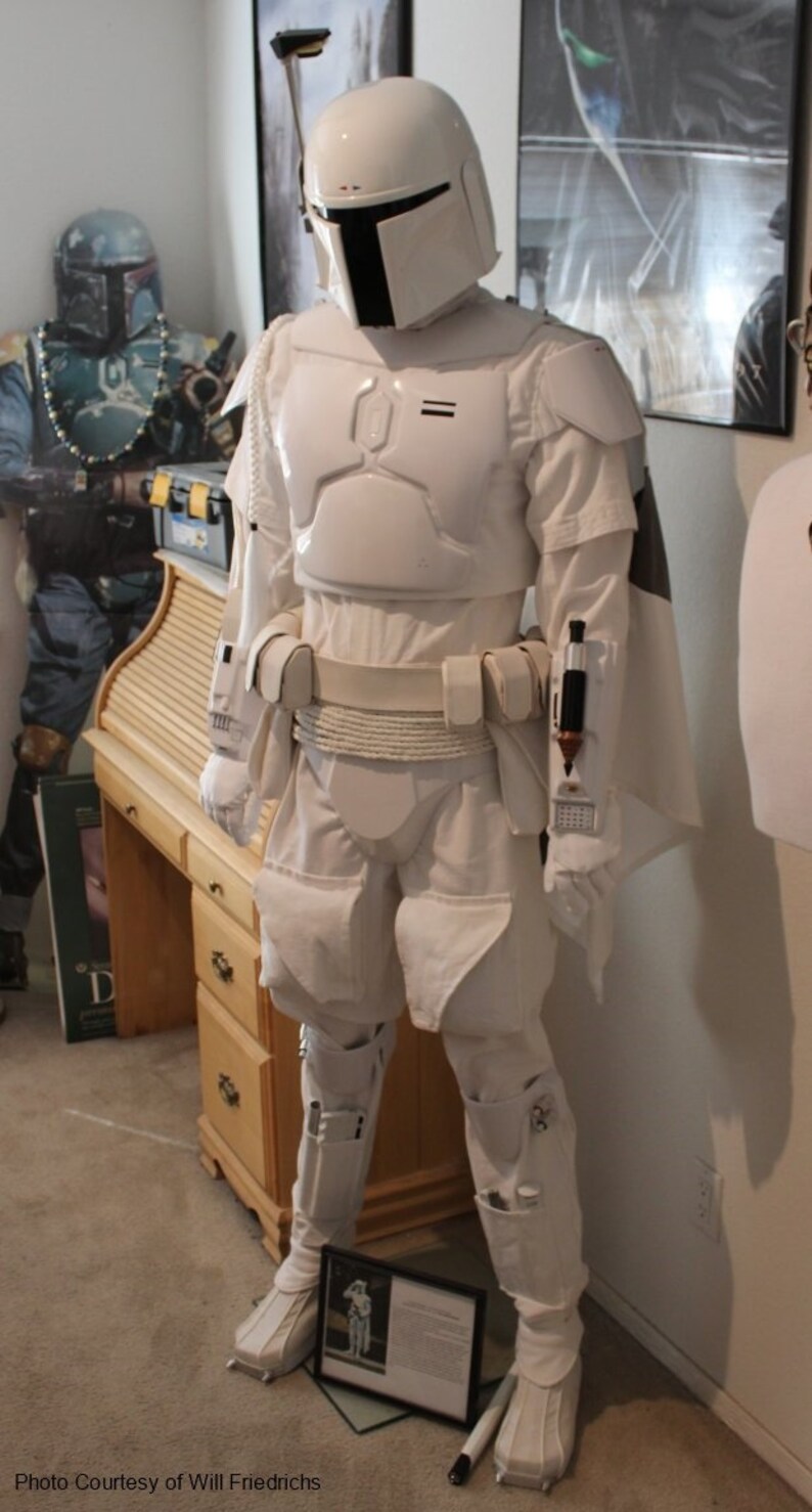 May include: A white Boba Fett costume with a white helmet and a white cape. The costume is standing on a brown carpet in front of a wooden desk. The costume is wearing a white belt with a rope detail. The costume has a white chest piece with a black visor. The costume has a white cape with a black trim. The costume has a white belt with a rope detail. The costume has a white chest piece with a black visor. The costume has a white cape with a black trim.