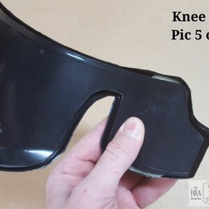 May include: Close-up of a black knee brace with a cutout for the kneecap. The brace is made of a flexible material and has a soft lining. The brace is being held by a hand. The text "Will Stitch" is visible on the brace.