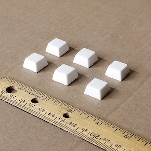 May include: Six white plastic keycaps for a mechanical keyboard. The keycaps are arranged in a staggered pattern on a brown surface. A wooden ruler is visible in the bottom left corner of the image.