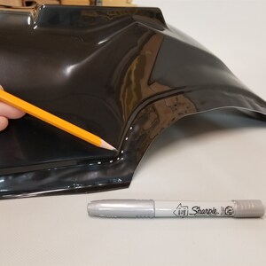 Puede incluir: Una pieza de pl&aacute;stico negro con una fila de puntos blancos a lo largo del borde. Una persona est&aacute; usando un l&aacute;piz amarillo para trazar los puntos. Un marcador plateado Sharpie est&aacute; sobre la superficie.