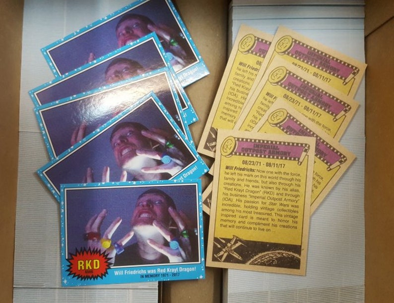 May include: A stack of yellow and brown trading cards with a blue and white card on top. The card features a man wearing a blue shirt and holding a ring. The text on the card reads "Will Friedrichs was Red Krayt Dragon! In Memory 1971-2017". The text on the yellow and brown cards reads "Imperial Outpost Armory 08/23/71 - 08/11/17".