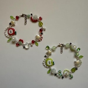 Puede incluir: Dos pulseras de cuentas. Una presenta dijes de manzana roja, cuentas rojas, estrellas blancas y detalles de hojas verdes. La otra pulsera tiene dijes de manzana verde, estrellas blancas y cuentas verdes y blancas. Ambas tienen cierres plateados.