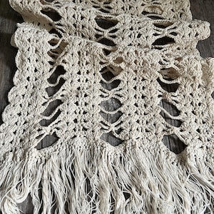 May include: An off-white crochet table runner featuring an intricate openwork pattern of repeating shell-like clusters and elongated vertical gaps. The runner is finished with long, loose, untwisted fringe tassels along the bottom edge.