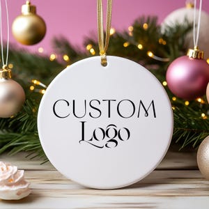 May include: White ceramic Christmas ornament with a black inscription that reads "CUSTOM Logo". The ornament is hanging from a gold ribbon against a pink background.