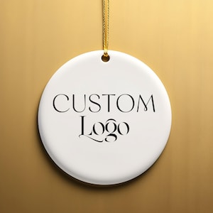 May include: White ceramic ornament with a gold string for hanging. The ornament has black text that reads "CUSTOM Logo".
