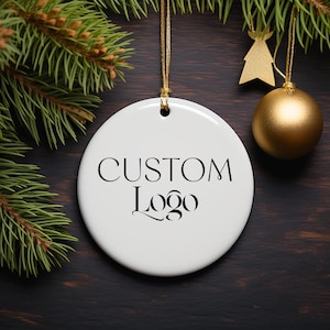 May include: White ceramic Christmas ornament with a gold string for hanging. The ornament has the text "CUSTOM Logo" printed in black.