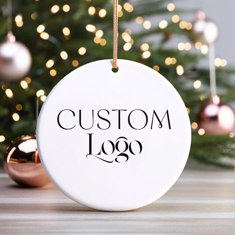 May include: White round ceramic ornament with a black inscription that reads "CUSTOM Logo". The ornament is hanging from a gold string against a blurred background of a Christmas tree with lights.